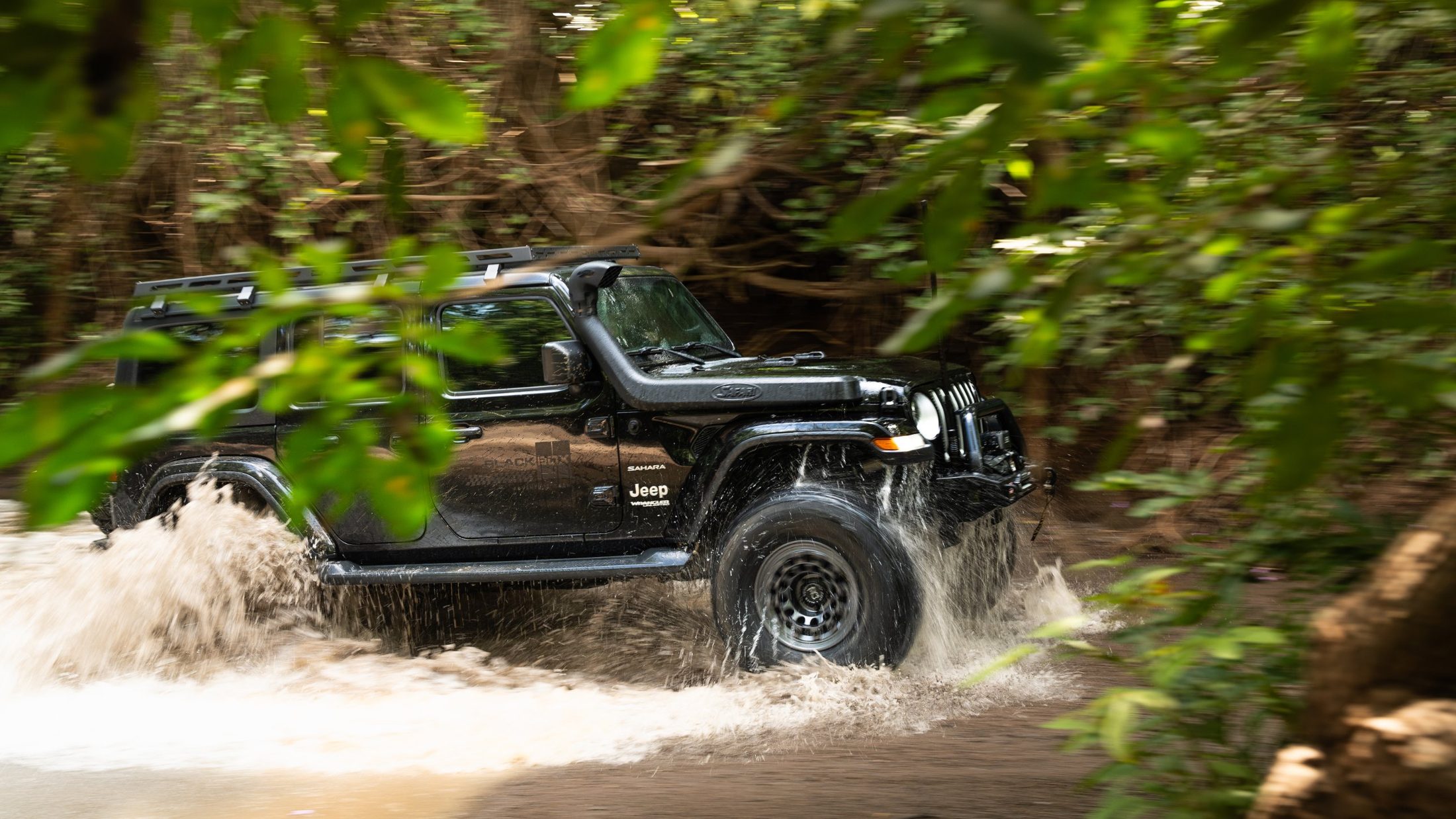 Black Jeep Wrangler splashing through a river, framed by leaves, photographed by Maxime Hugo