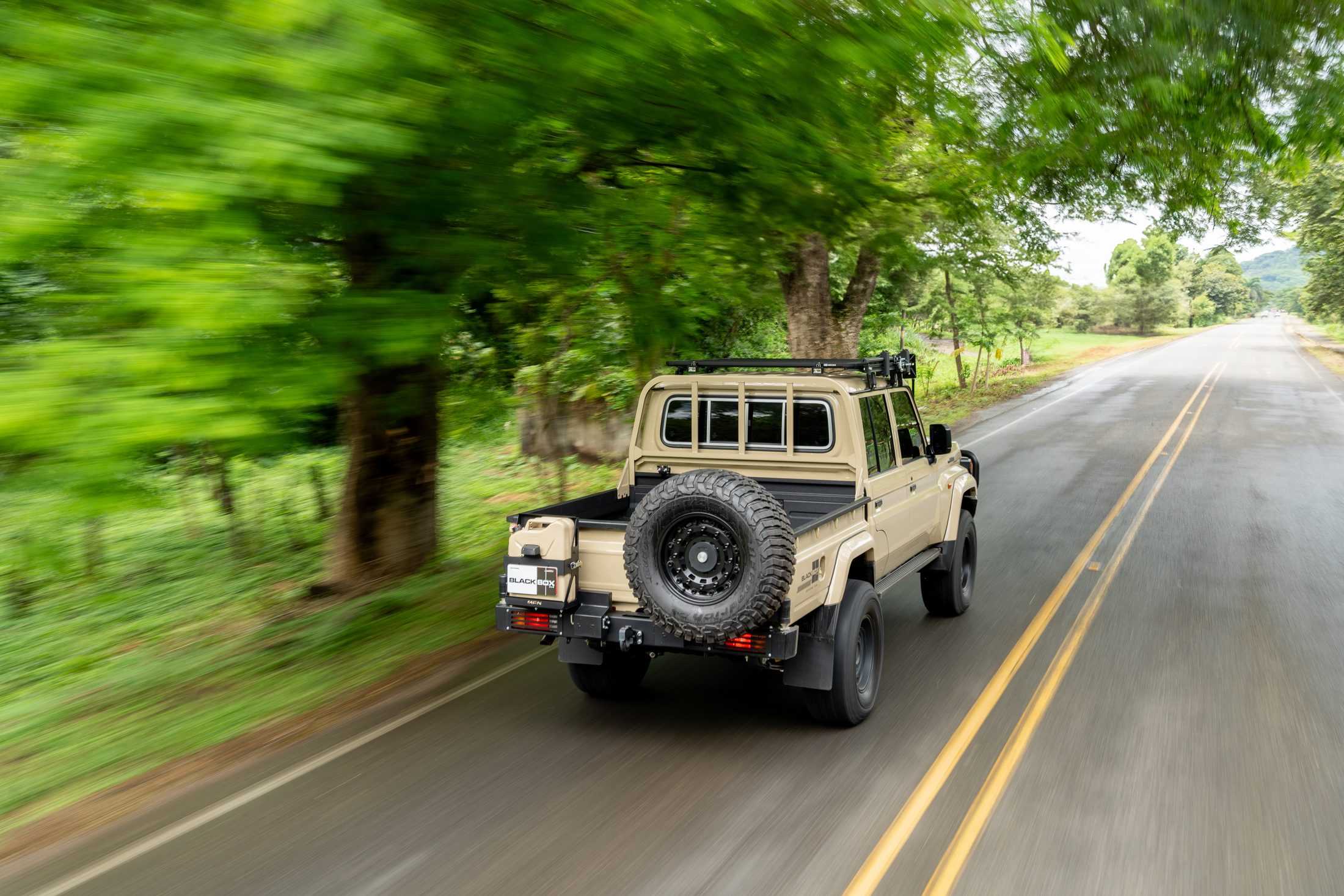 Beige Land Cruiser 70 pickup seen from behind on a tree-lined road, photographed by Maxime Hugo